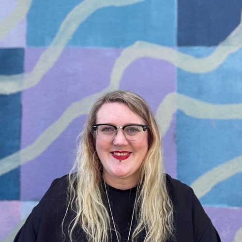Rhen smiles into the camera while standing in front of a multicoloured background. They have long blonde hair and are wearing glasses and red lipstick.
