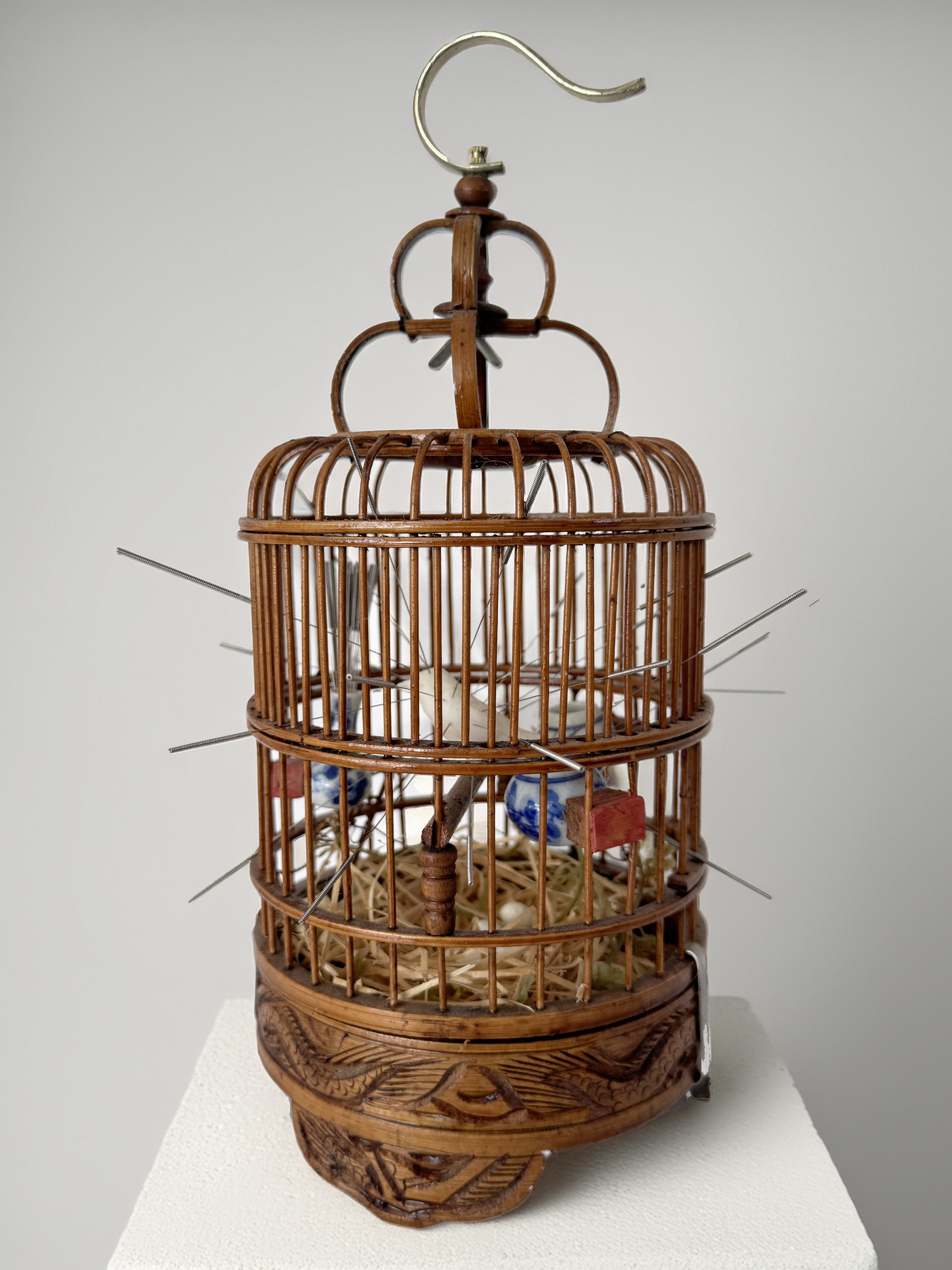 A wooden bird cage with a bird and various objects in it and lots of skewers sticking out of it