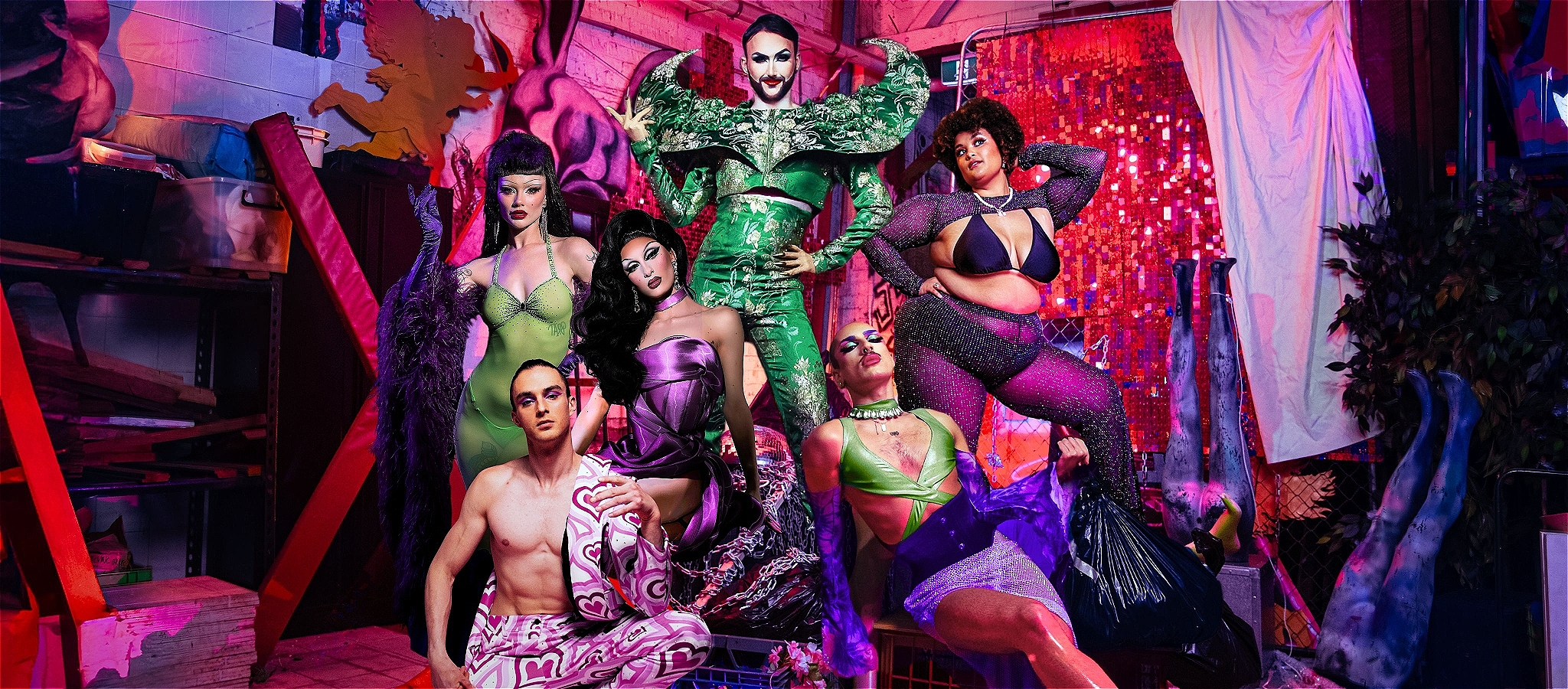 Eight performers in colourful and glittering costumes pose together in a warehouse, surrounded by props, neon lights, and sequinned backdrops.
