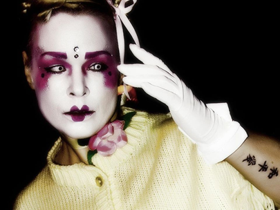 Androgynous person wearing lemon cardigan and white gloves, pale makeup, heavily shaded eyes and mouth