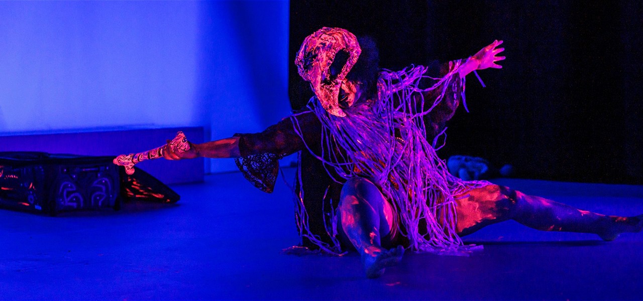 A person wearing UV body paint crouches on the ground and holds out a bone prop.