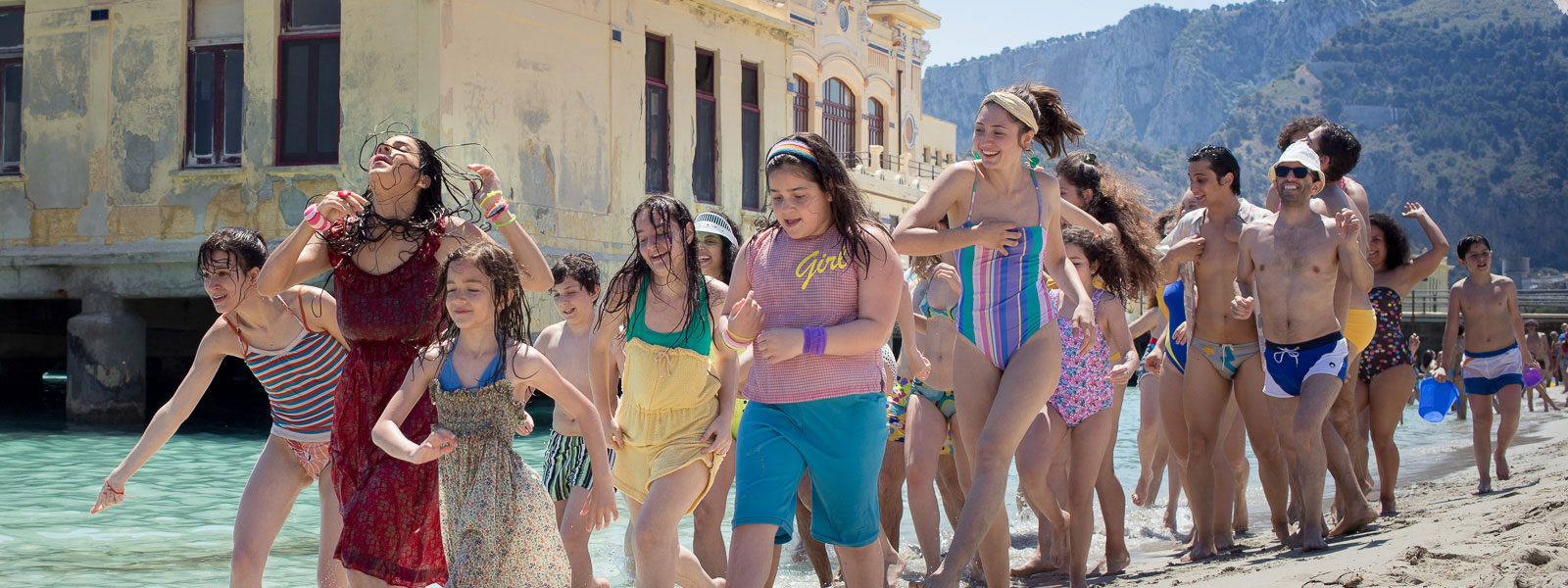 Lots of people in beach attire following a person in a scarlet dress in a seaside setting