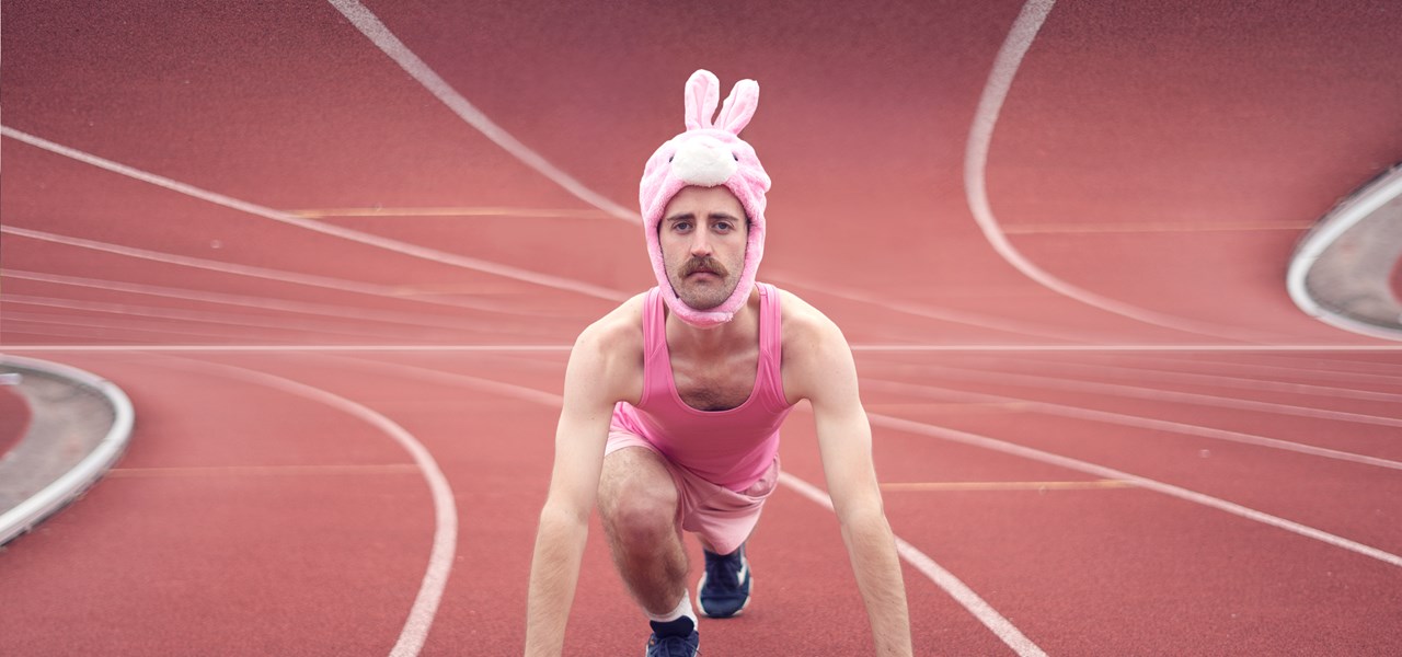 A man wearing a pink bunny hat and pink sportswear positioned in a standing sprint stance on a running track