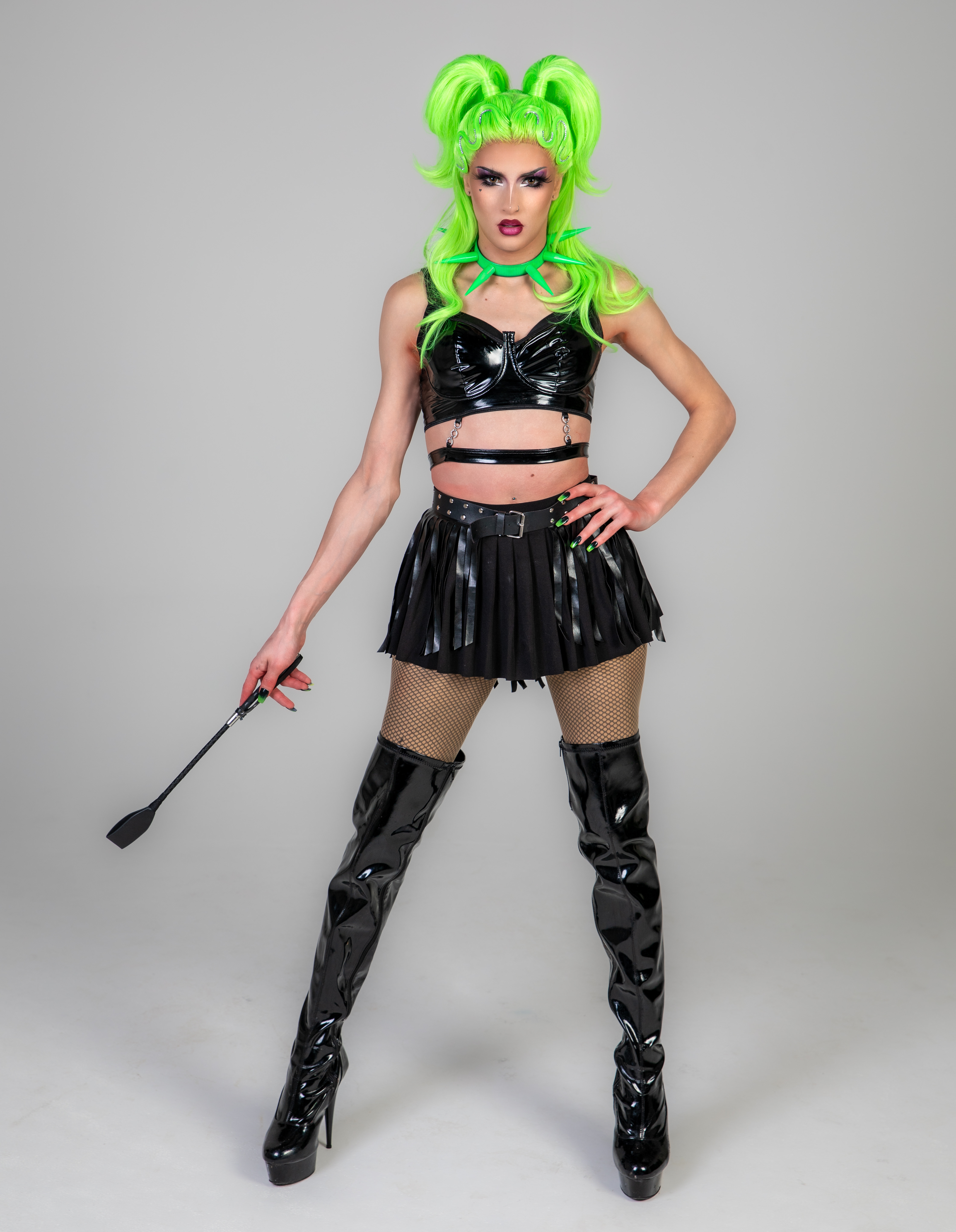 Clepto Nymph dressed in green wig and black leather with one hand on their hip and the other holding a riding stock