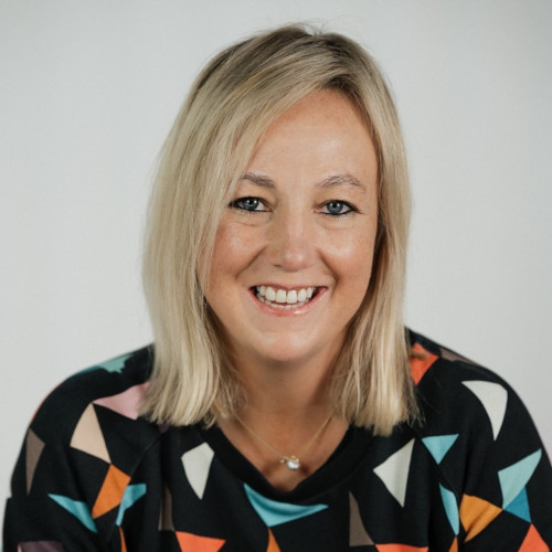 Headshot of Kelly smiling. Kelly has long blonde hair and is wearing a black top covered with different coloured triangles.