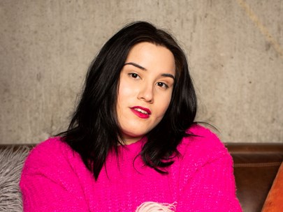 Sarah poses with their head on a slight angle and wears bright red lipstick. They have black, silky shoulder length hair, dark eyes and pale skin. They are wearing a hot pink jumper.