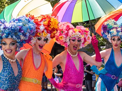 Four colourfully dressed drag queens, with rainbow umbrellas in the background