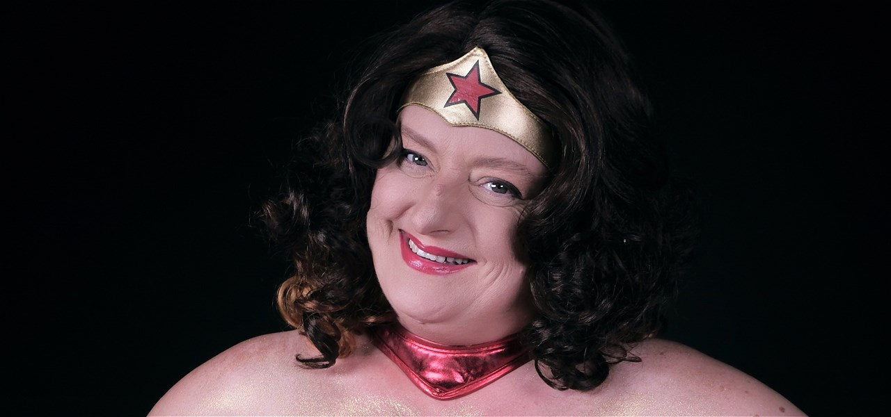 A smiling woman dressed up as wonder woman looking directly at the camera. She is white and has curly black and brown hair that is shoulder length.