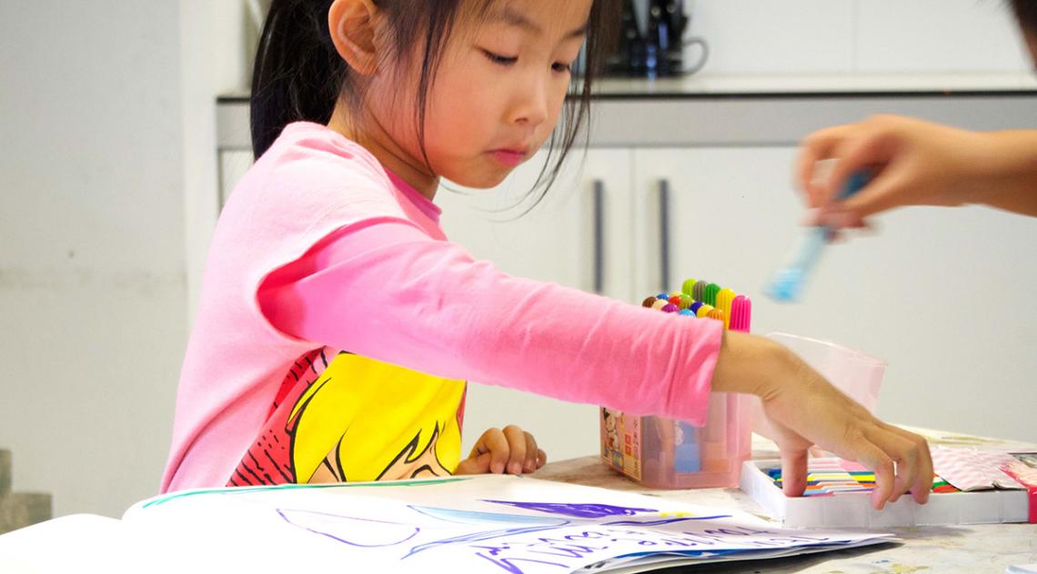 Young Asian girl reaching for pencils, artwork spread out on table in front of her