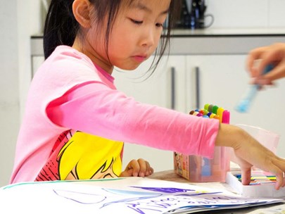Young Asian girl reaching for pencils, artwork spread out on table in front of her