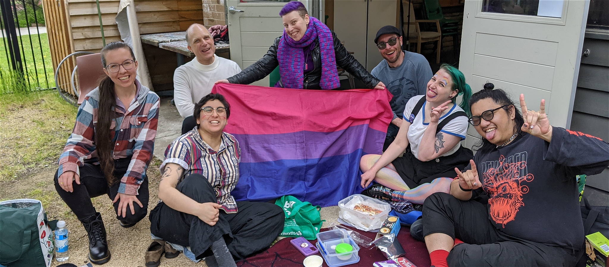 A group of smiling people seated around a picnic blanket, with a bisexual flag held up between them. The people are pulling silly faces and laughing.