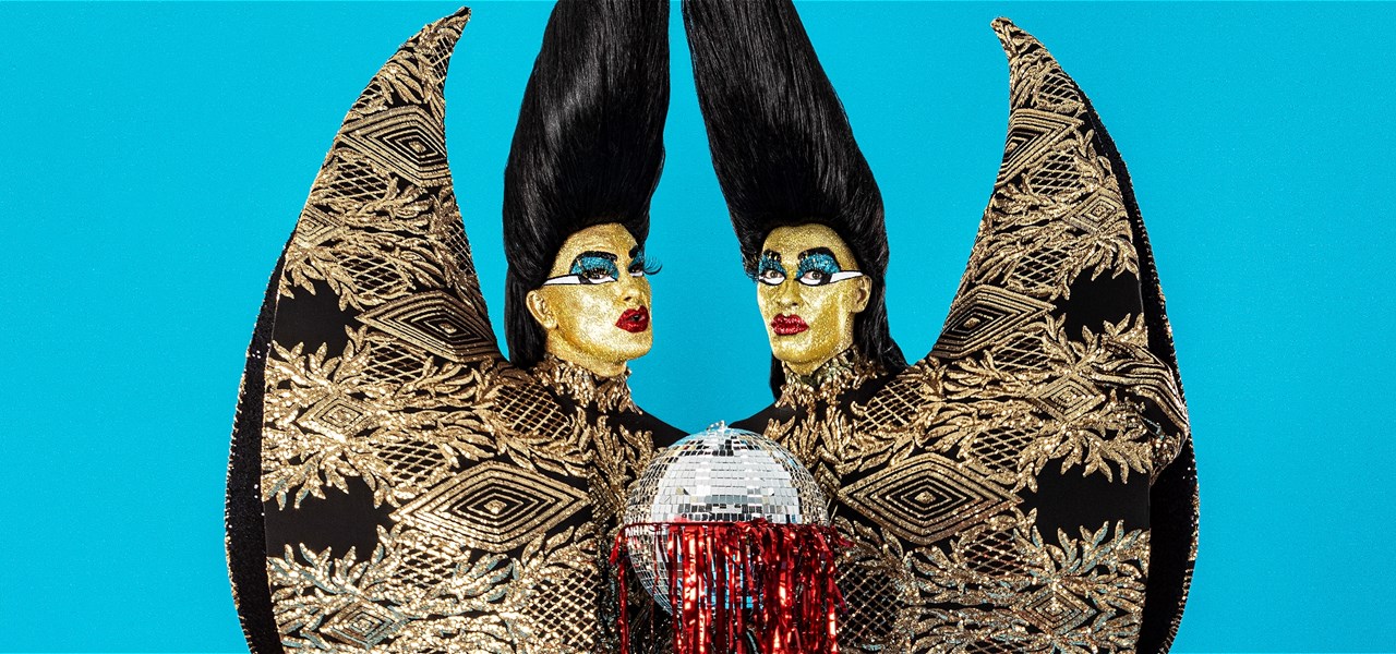 Two performers wearing tall black wigs and sparkling black and gold bodysuits, hold a silver ball dripping with red tinsel