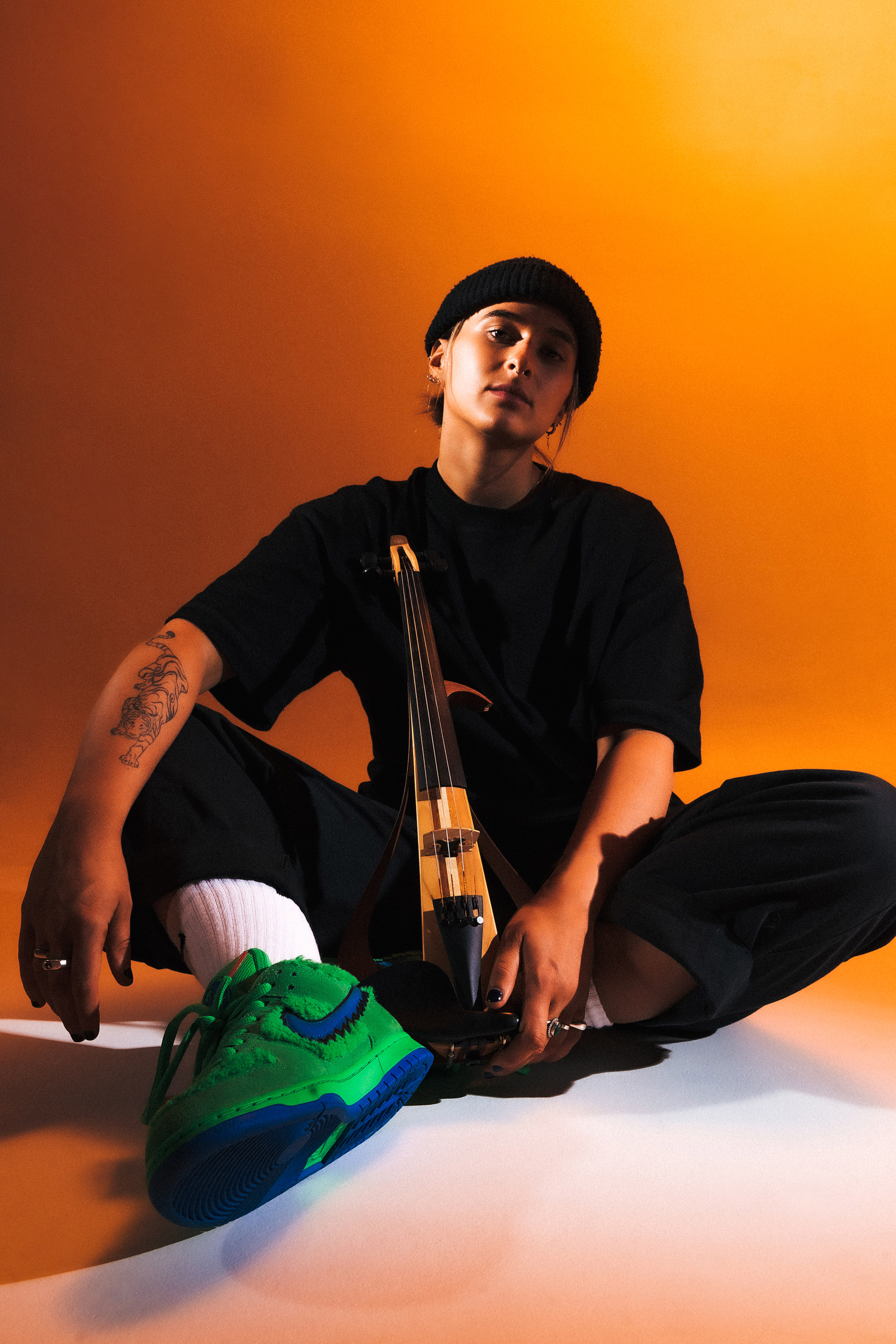 Umbra Moon holding a string instrument dressed in a black outfit including black beanie against orange lighting