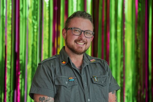 Headshot of Teague in a grey open-neck shirt, standing in front of a striped curtain