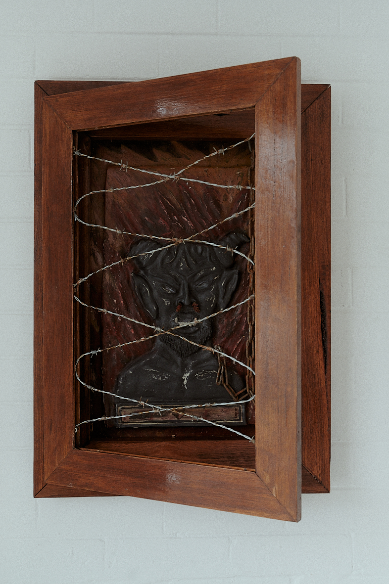 A dark portrait behind a wooden frame with barbed wire criss-crossing in front of the image