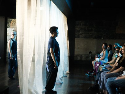 Photo from the side of a stage with one person in front of the curtain facing an audience, and somebody waiting behind the curtain