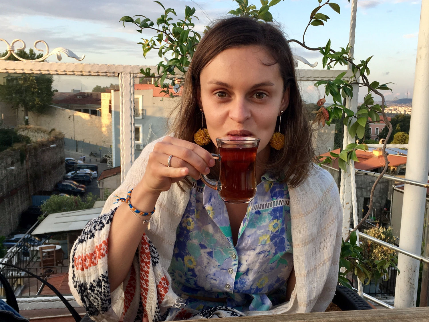 Jacqui sipping a tea while standing in an outdoor patio, with a (European looking) modern town in the background