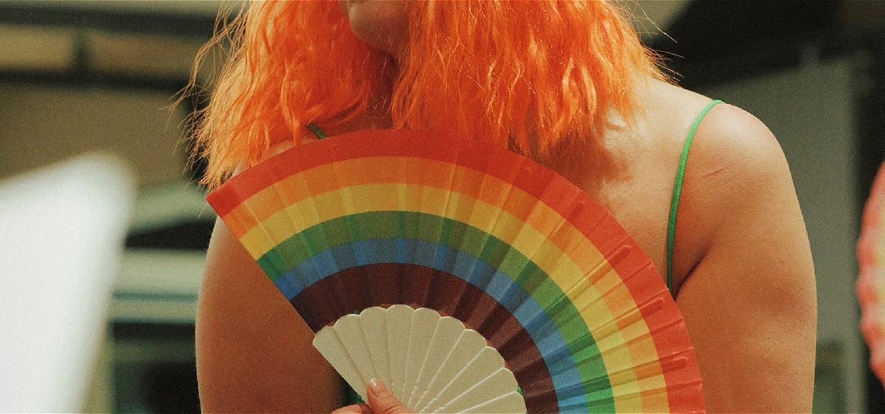 Image is of a person with orange hair holding a rainbow fan