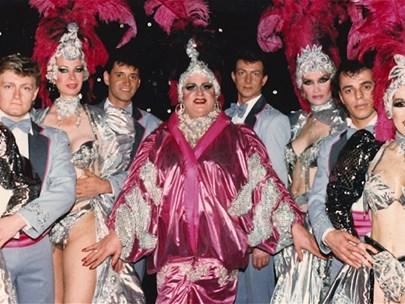 Eight performers dressed in blue suits and showgirl costumes stand around the main performer who is lavishly dressed in a pink robe and headdress.