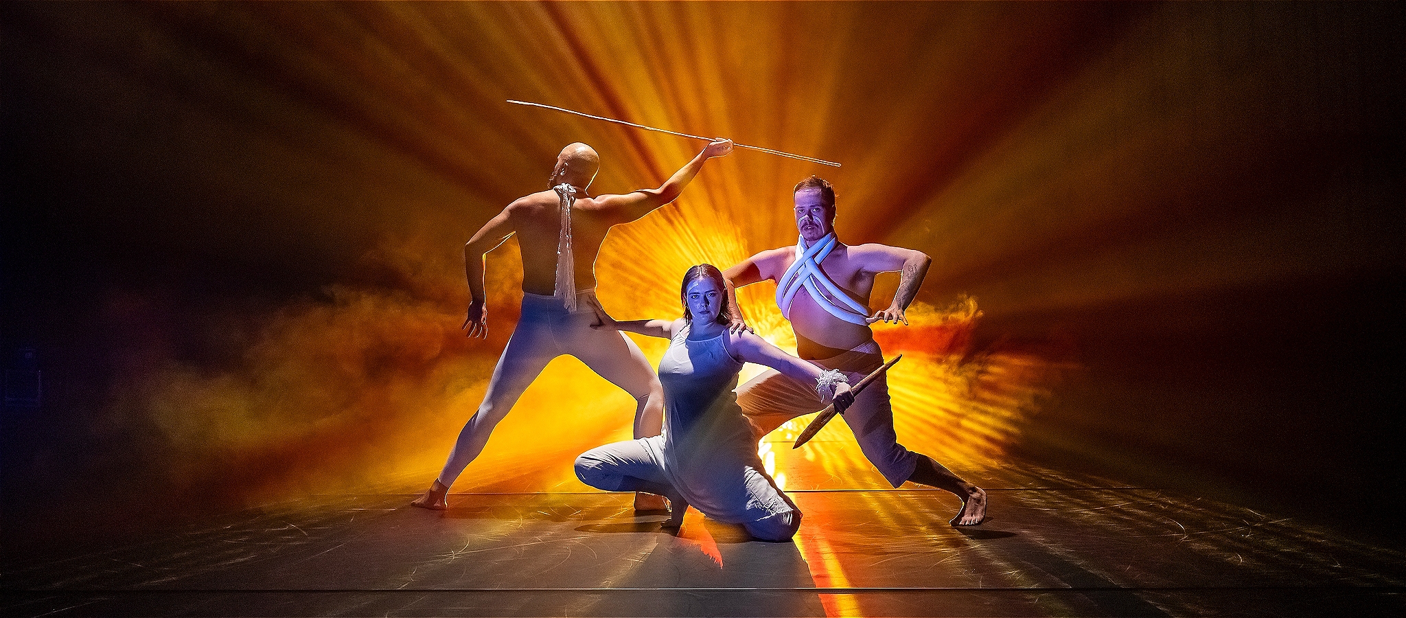 Three performers in strong poses, yellow and orange beams of light surround them.