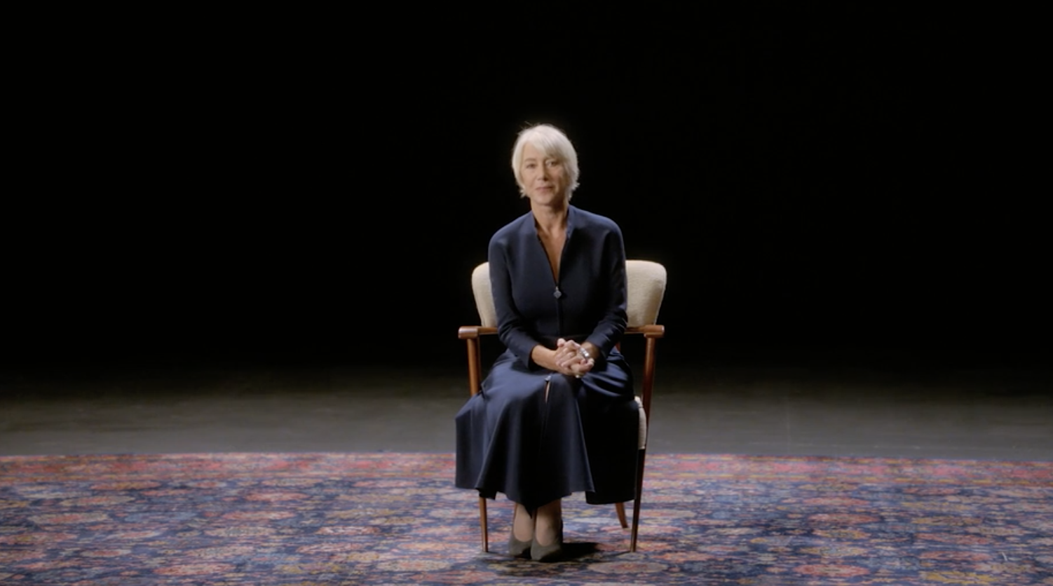 Actor Helen Mirren sitting in a chair, hands clasped together, in a large space