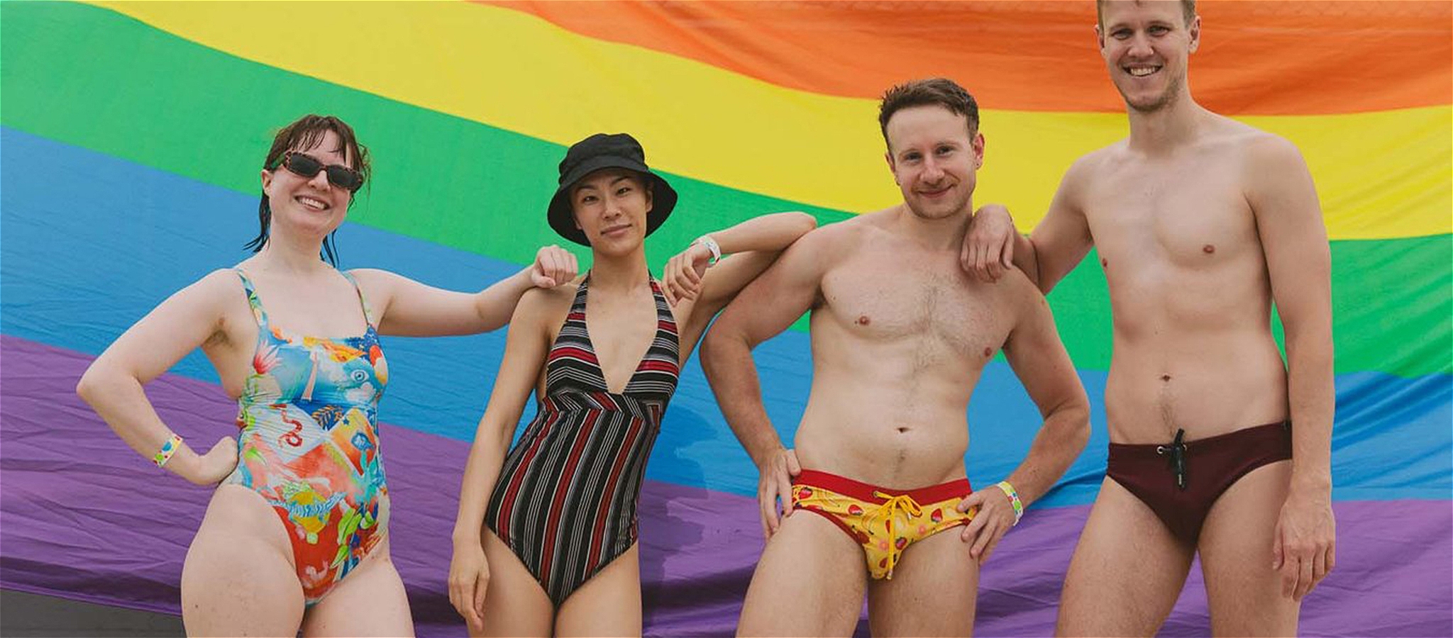 Four people pose in their bathers in front of a rainbow pride backdrop.