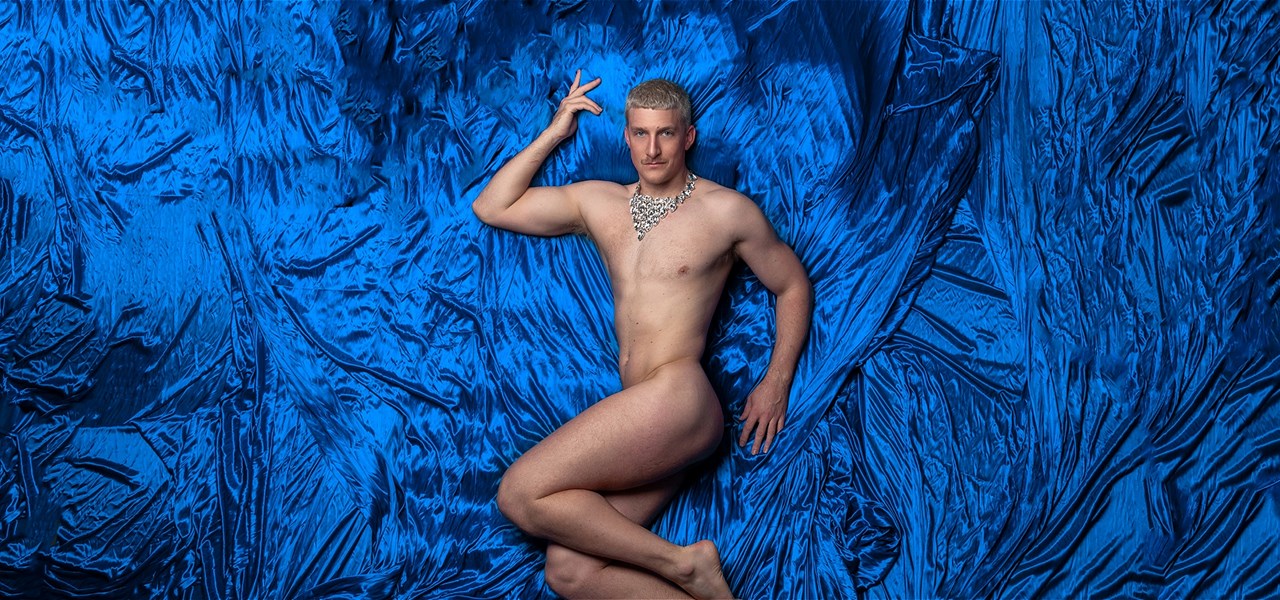 Ziegfeld Boy lies on a bed of royal blue silk sheets wearing nothing but a diamond necklace