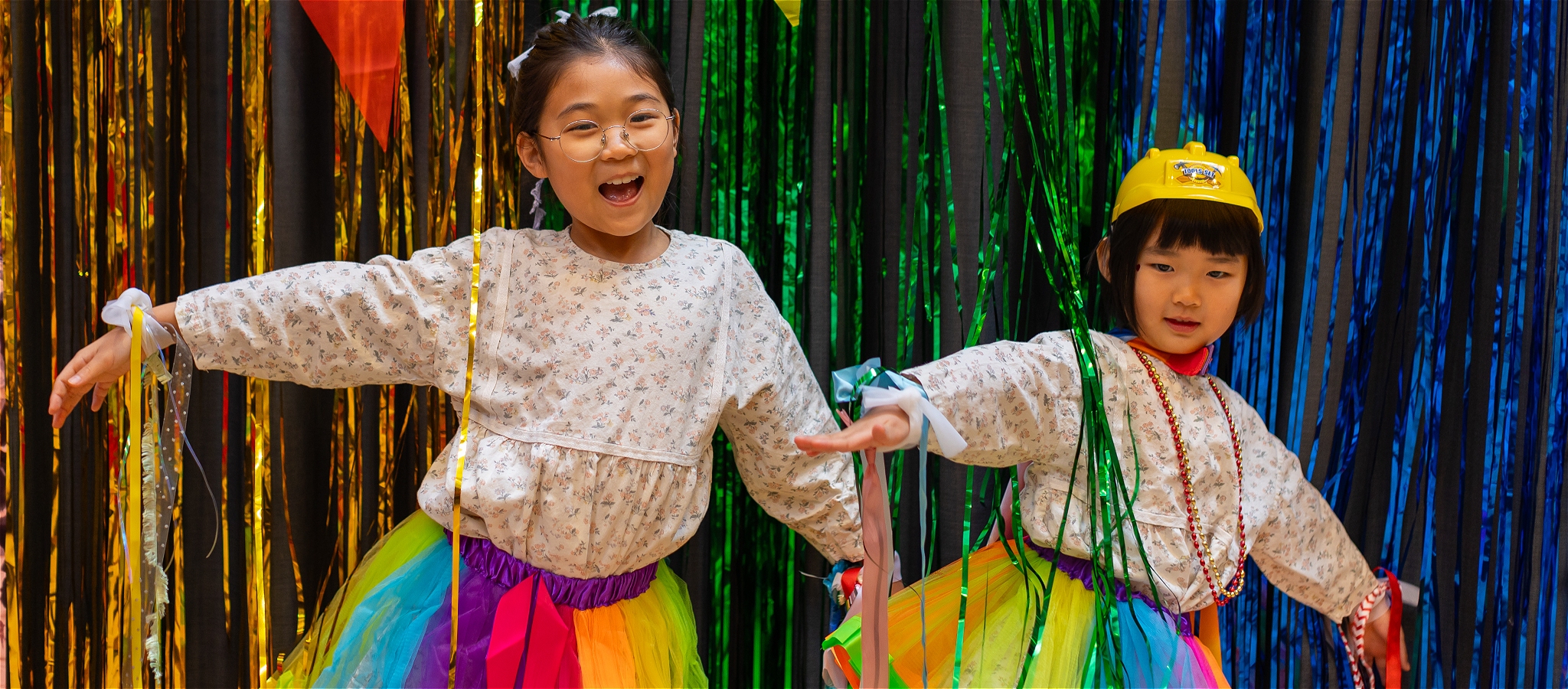 Two children wearing rainbow tutus and floral tops pose with arms stretch out with smiling facesinfront of a glitter curtain.
