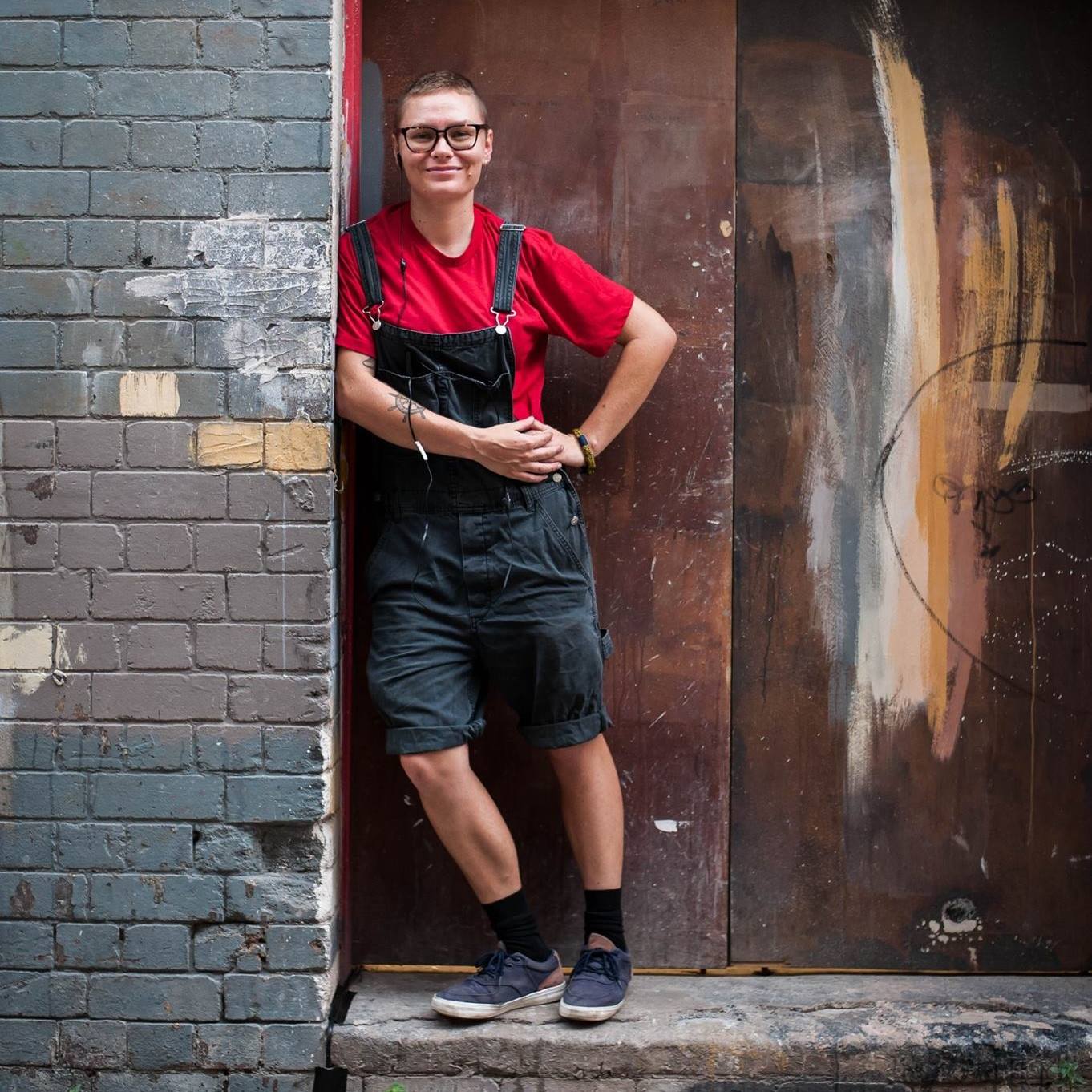 Mack dressed in overall shorts and red t-shirt leaning on a wall in a grungy building