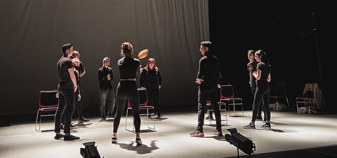 Group of 9 people standing in a circle wearing all-black passing a ball around on a stage