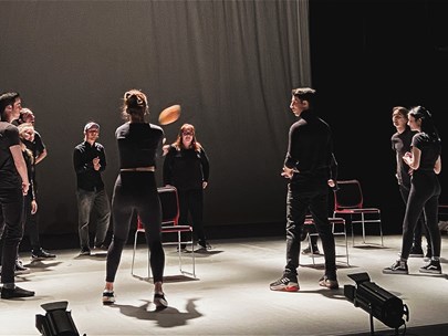 Group of 9 people standing in a circle wearing all-black passing a ball around on a stage