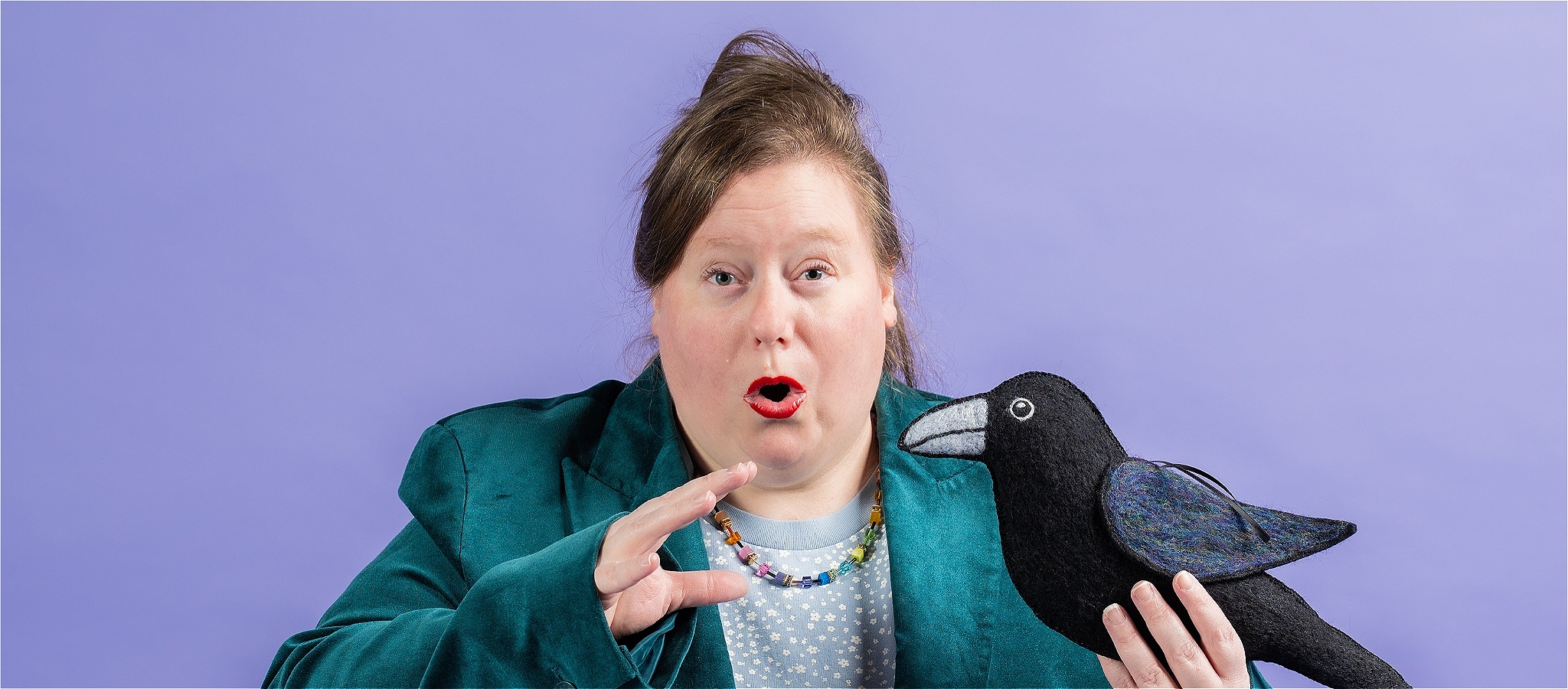 A woman, looking surprised, wearing a dark green blazer, blue tshirt and rainbow necklace. Holding a plush raven.