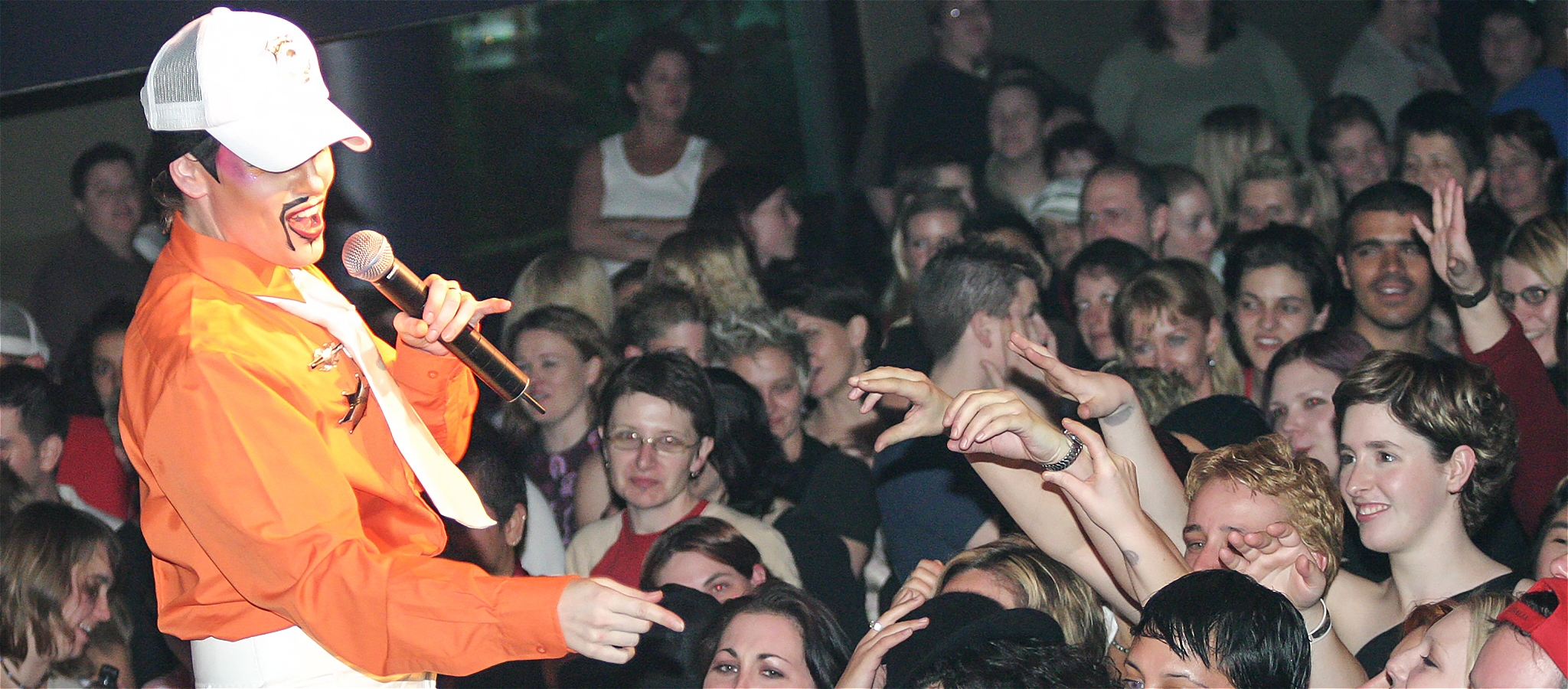 A performer wearing an orange shirt and a white cap in front of a large crowd