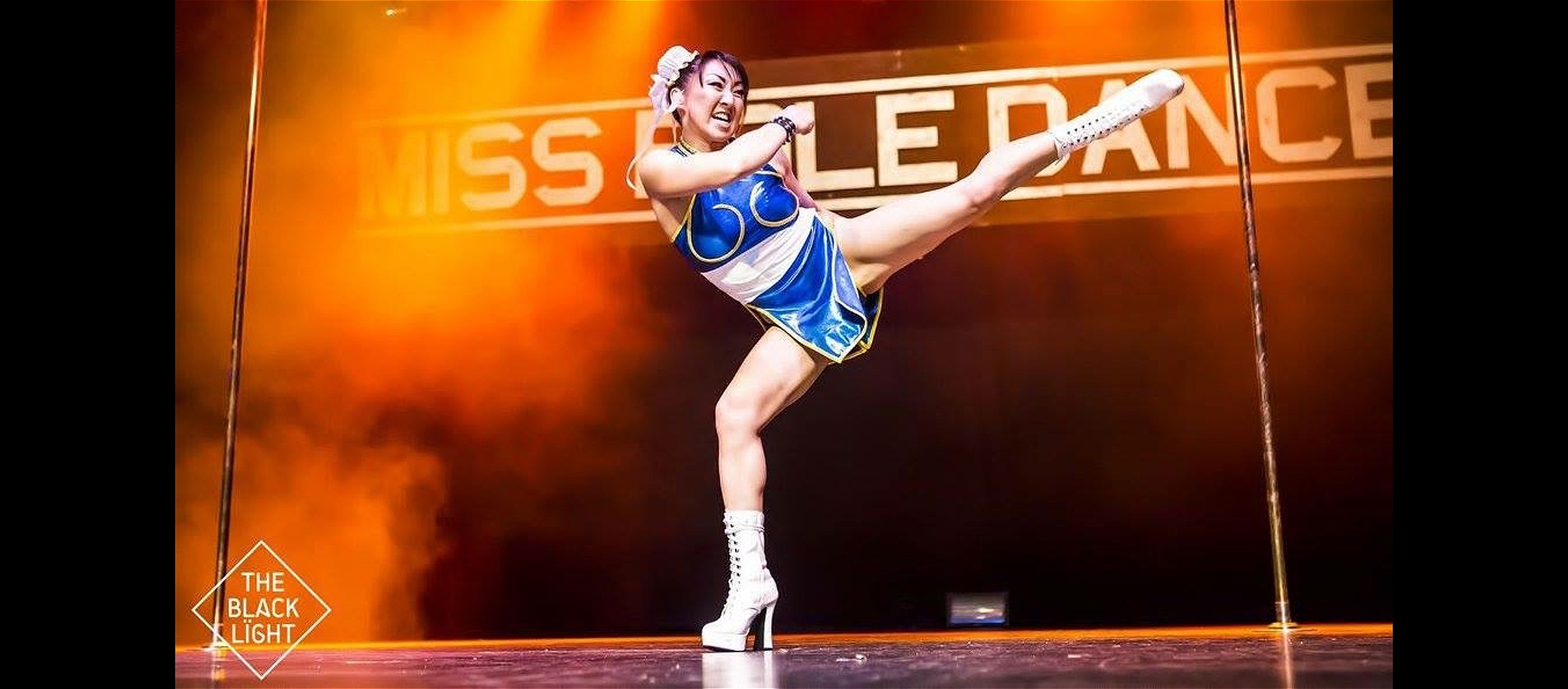 Asian woman dressed as Chun Li from Street Fighter executes a high side kick onstage with two poles