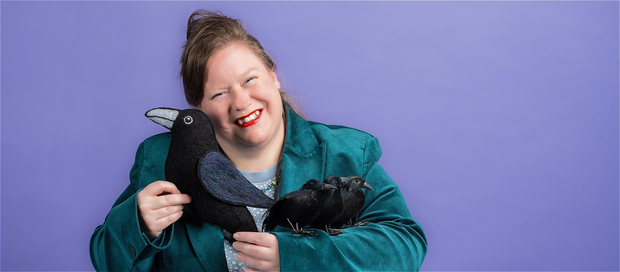 A smiling woman, wearing a dark green velvet blazer, blue tshirt and rainbow necklace, holding a large plush raven and three smaller toy ravens
