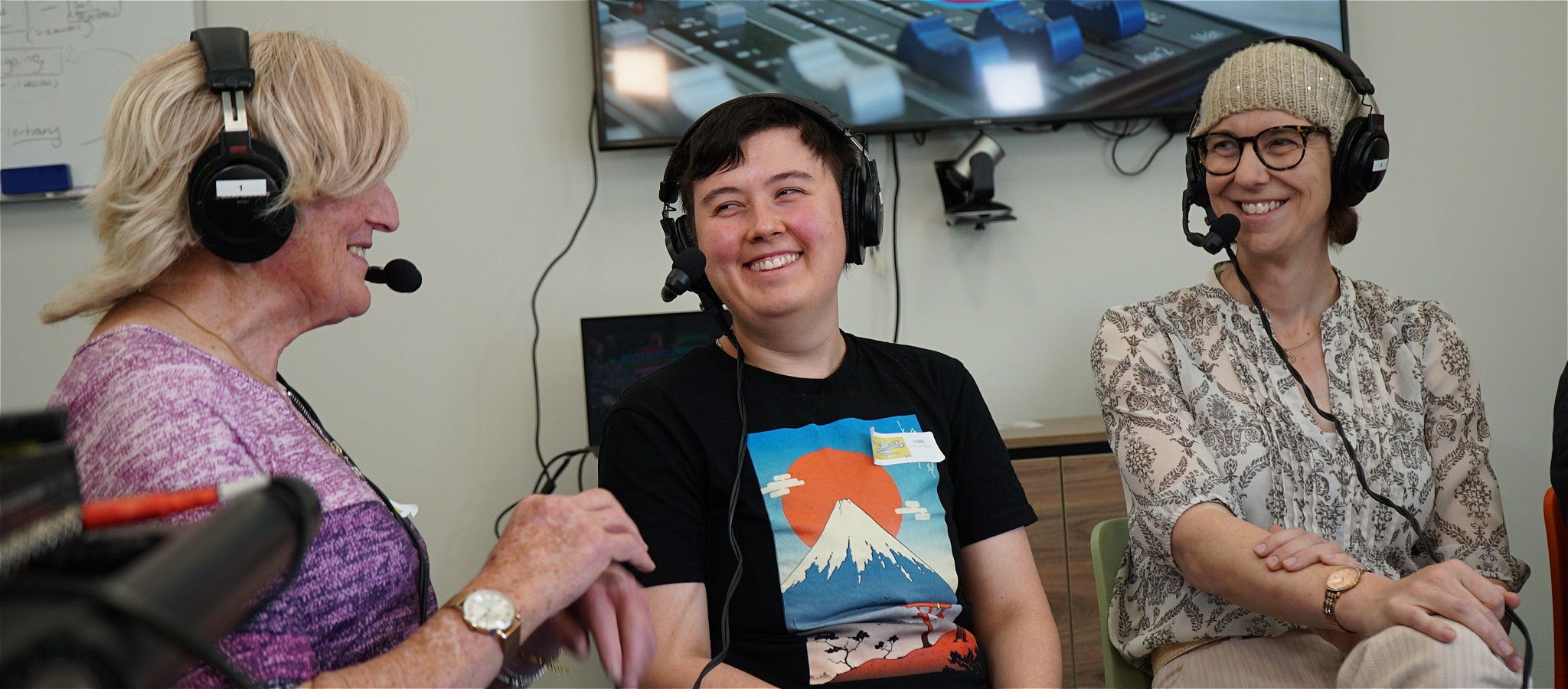 Three people wearing headphones with microphones, seated next to each other and smiling at one another.