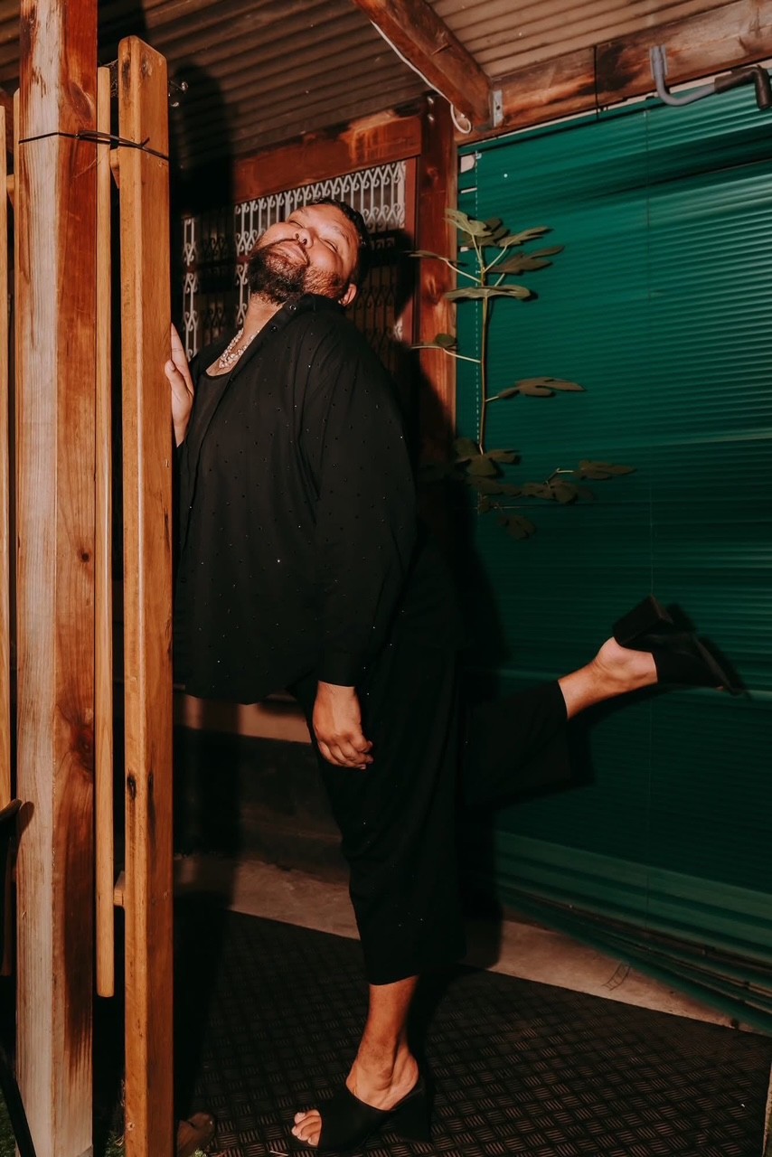 2Joocee dressed in black leaning against a door frame head tilted back and one leg raised