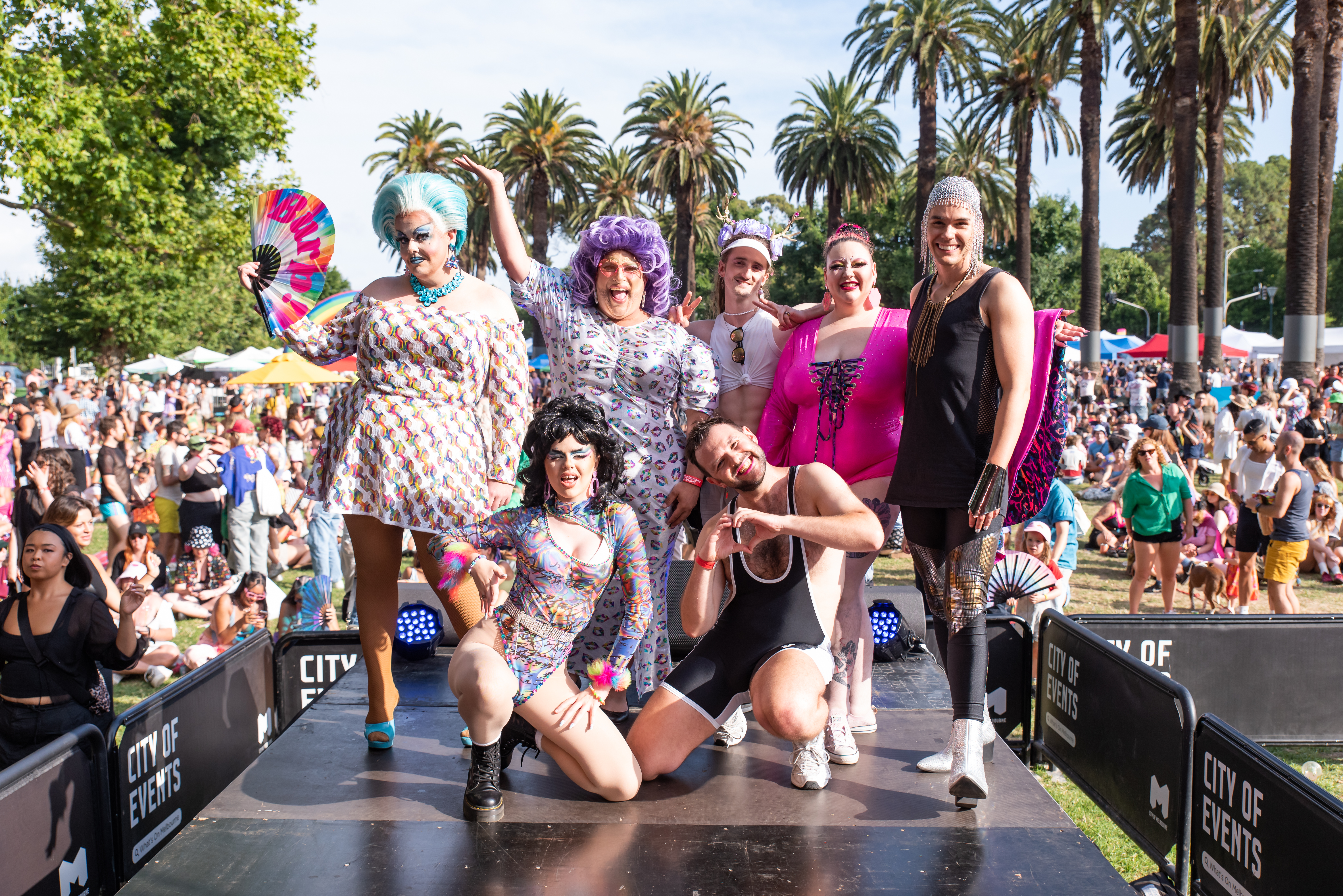 seven colourful performers on stage with a large crowd in the background
