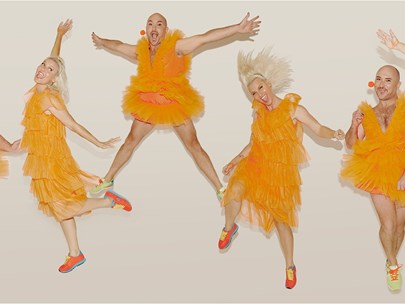 Six performers dressed in orange tulle dresses are in a row, smiling, dancing and jumping