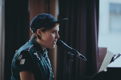 Kip speaks into a microphone. They have short brown hair and are wearing a baseball cap and a spiked vest over a t-shirt.
