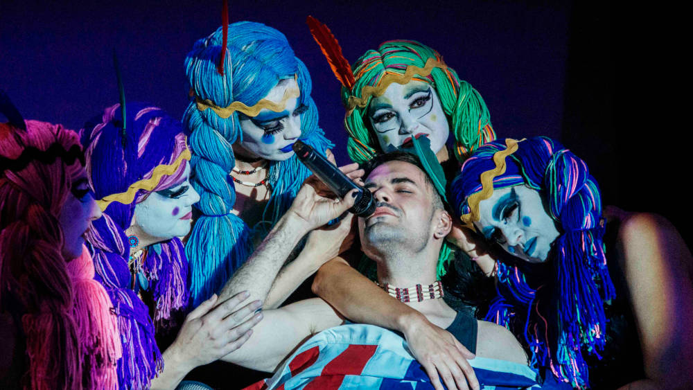 Man singing into mic, surrounded by three people with white-painted faces, holding their arms around him