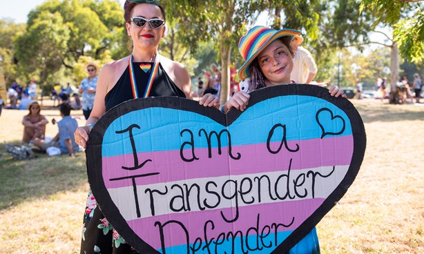 Two people holding a heart-shaped banner: "I am a Transgender Defender"