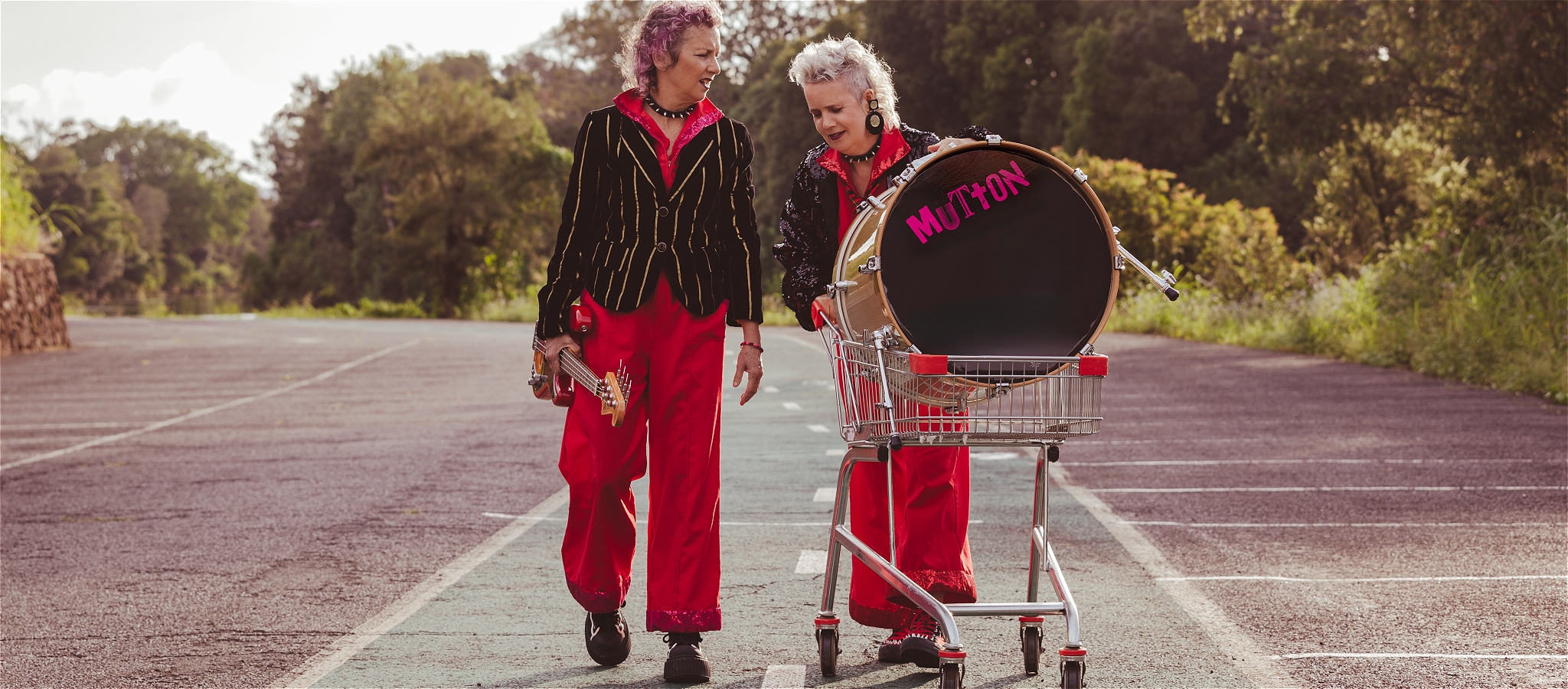 Two women in red jumpsuits push a trolley with a drum in it, talking, in a carpark. One carries a red bass guitar.