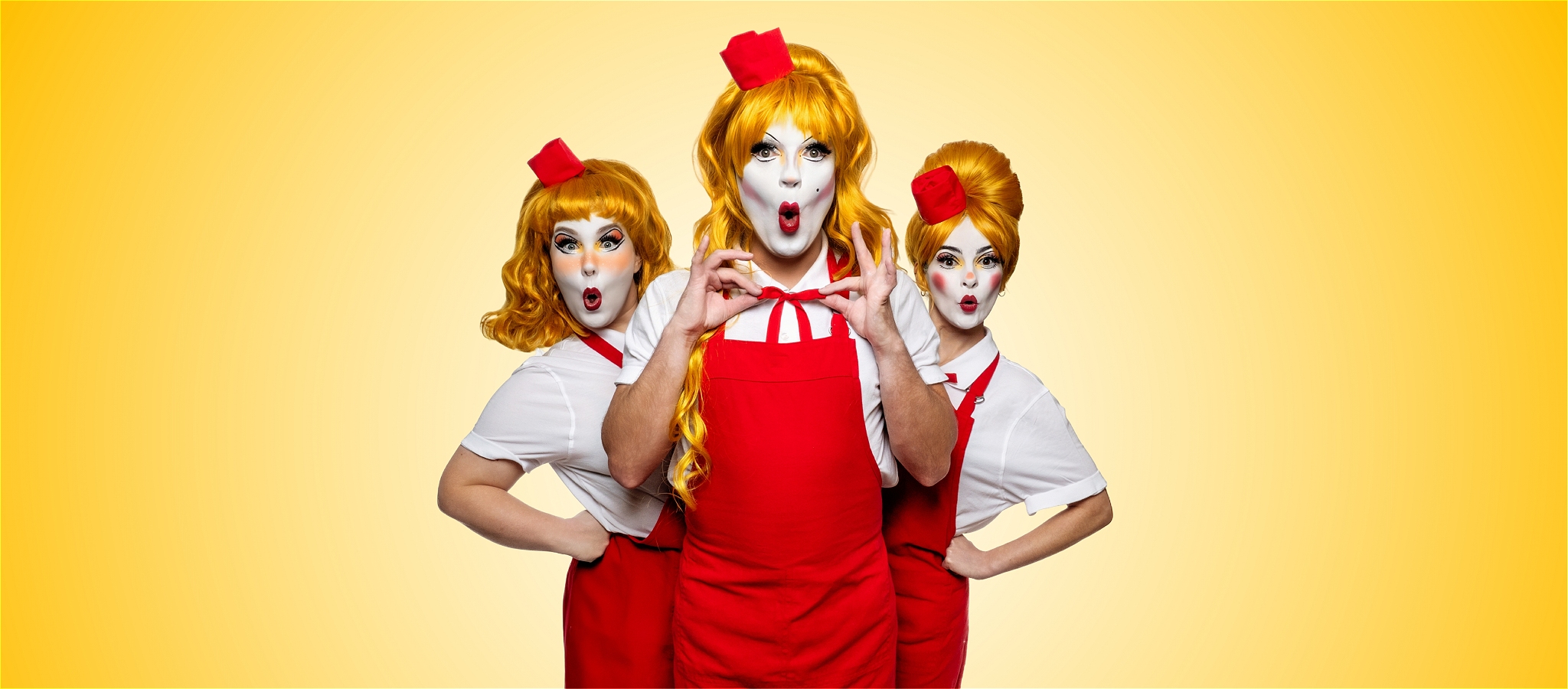 Three people wearing red aprons and soda jerk caps with yellow wigs looking surprised.