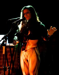 Sophie Rose standing on stage in front of two microphones, wearing orange slacks and black top