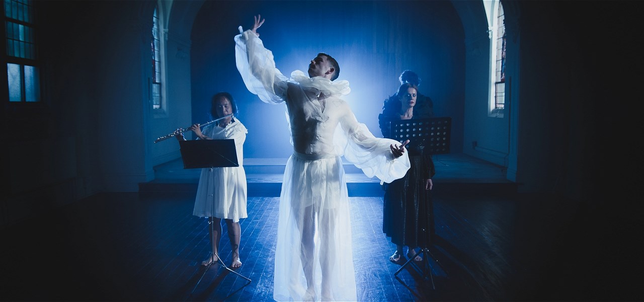 4 people are pictured in a dark church back lit with a blue-white light. One person is featured in the centre in a white outfit and they are dancing.