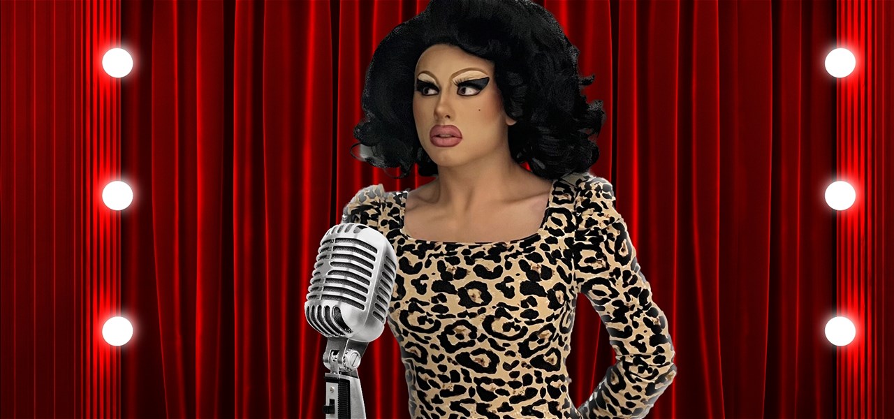 Drag queen Fanny Adams, wearing a black wig and leopard print dress, stands behind a microphone.