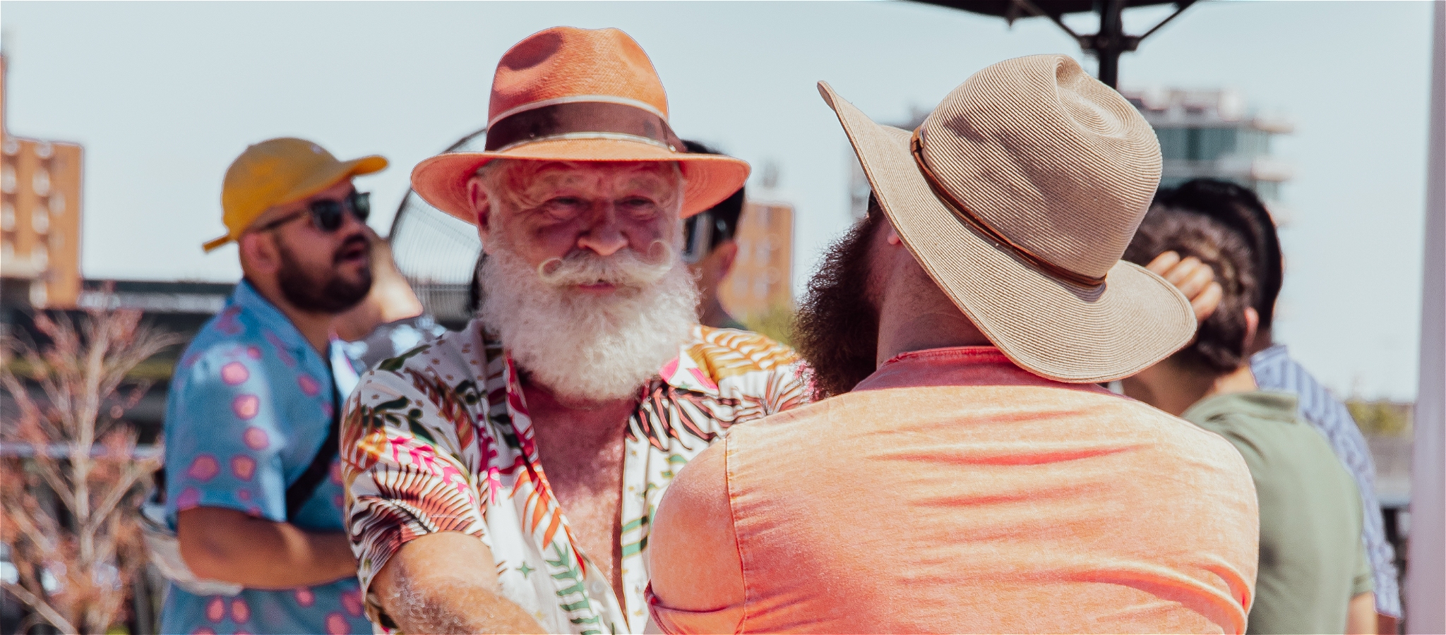 Two older people dancing and holding hands together outdoors in the sunshine, wearing vibrant casual clothes and sun hats.