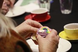 Two hands holding a rainbow-decorated cup cake; cups of tea in the background