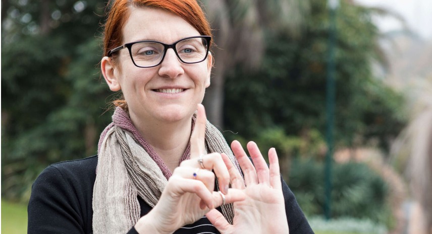 A person with red hair and black glasses is standing in a park and signing